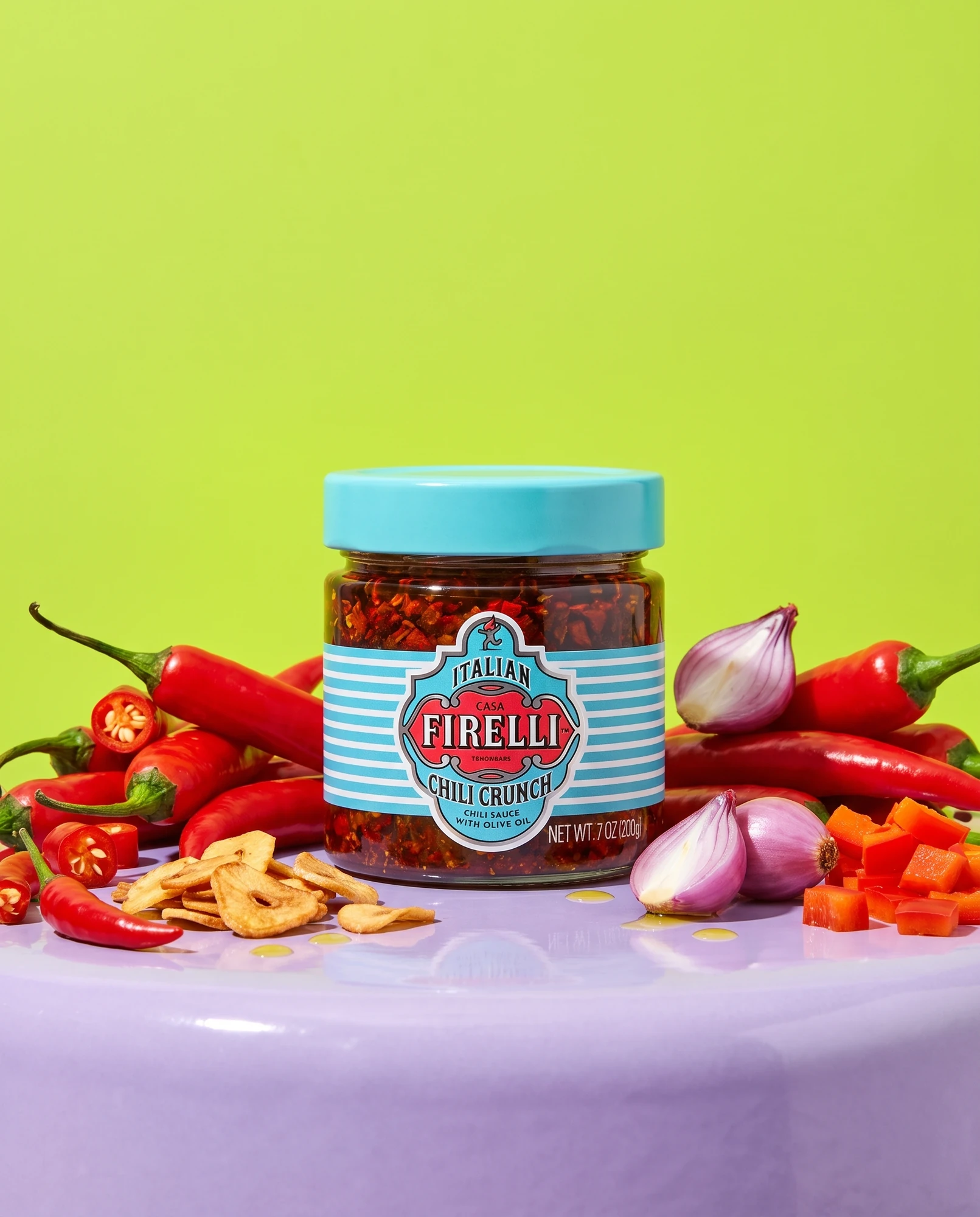 Vasetto di Chili Crunch Casa Firelli tra peperoncini freschi, chips di aglio e cipolla su sfondo verde lime