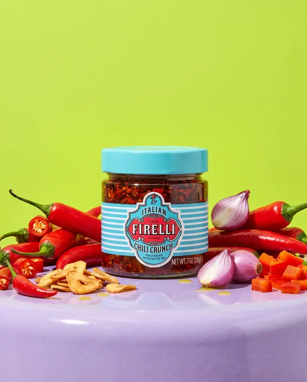 Vasetto di Chili Crunch Casa Firelli tra peperoncini freschi, chips di aglio e cipolla su sfondo verde lime