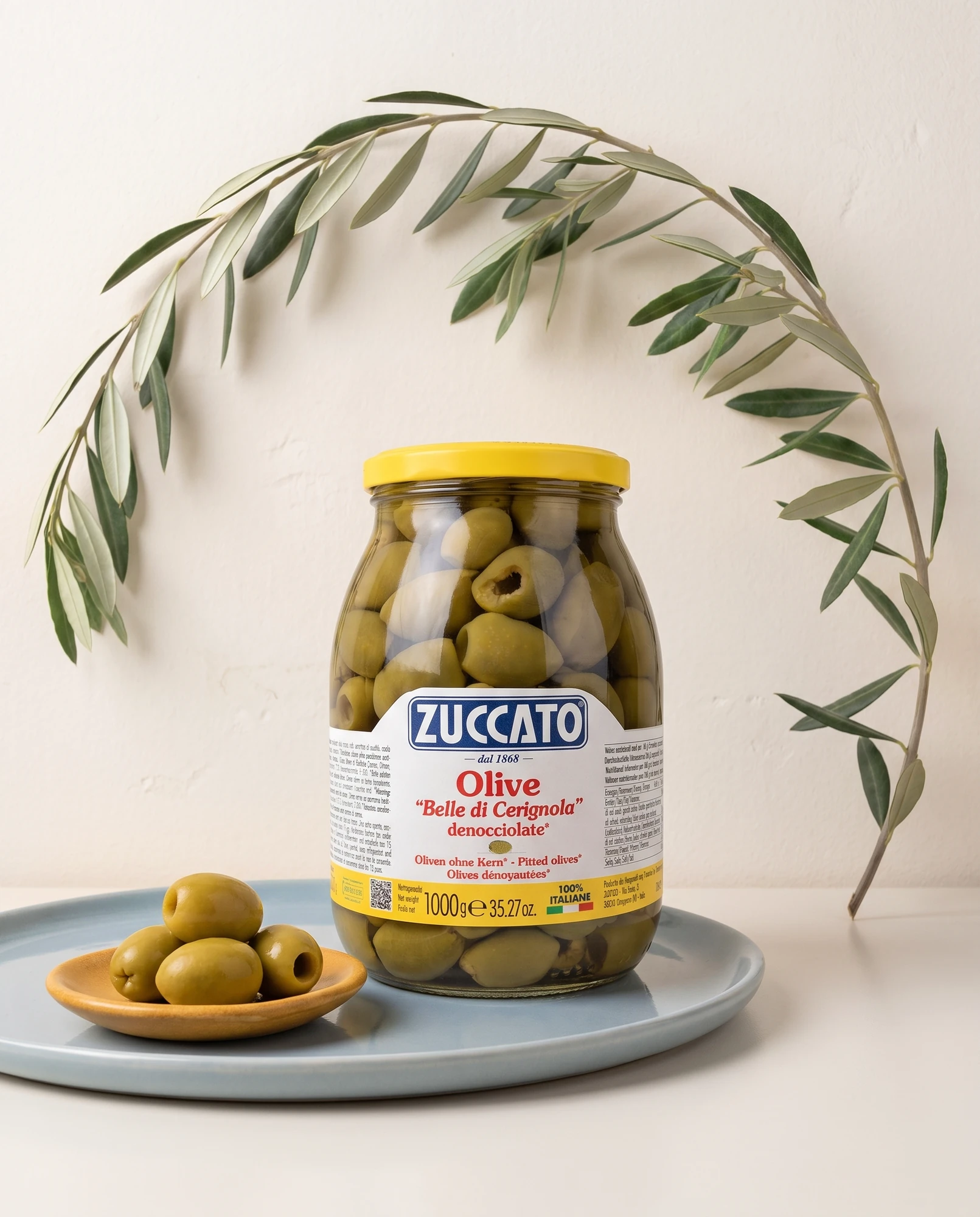 Barattolo di olive Belle di Cerignola Zuccato con un ramo d'ulivo e una ciotola di olive su piatto azzurro